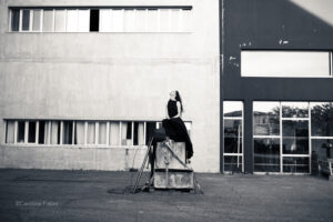 Flore, artiste de la compagnie d'ielles est perchée sur des blocs de pierre, habillées tout de noir, silhouette de sirène tragique dans un décor béton.